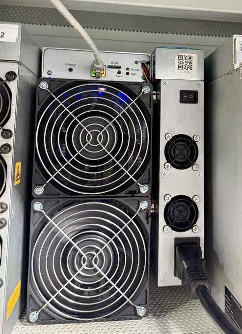 The back panel of a cryptocurrency mining device showing two large cooling fans with protective grills, various ports, and indicator LEDs. An ethernet cable and power cable are connected to the unit.