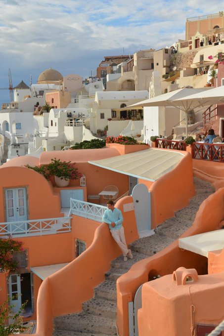 A scenic view of Oia, Santorini, Greece, showing white and terracotta-colored buildings with a dome, where a person in a turquoise jacket and beige pants leans against a peach-colored wall on a stone pathway.