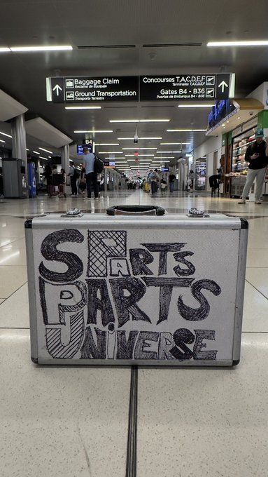 A silver briefcase with 'SPORTS PARTS UNIVERSE' handwritten in black marker sits on the floor in an airport terminal, with signs for Baggage Claim, Ground Transportation, and Concourses visible in the background.
