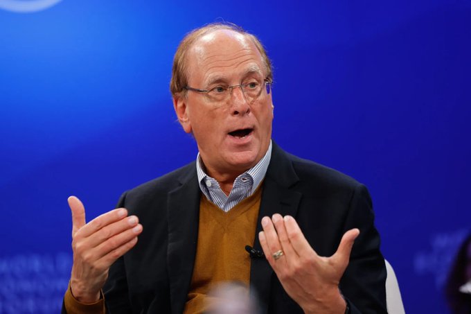 An older man with glasses wearing a dark blazer over a brown sweater and checkered shirt, speaking while gesturing with both hands against a blue background.