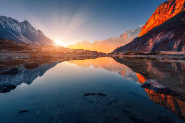 A scenic mountain landscape at sunset with snow-capped peaks reflected in a calm lake, with golden sunlight illuminating the rocky cliffs on the right side.
