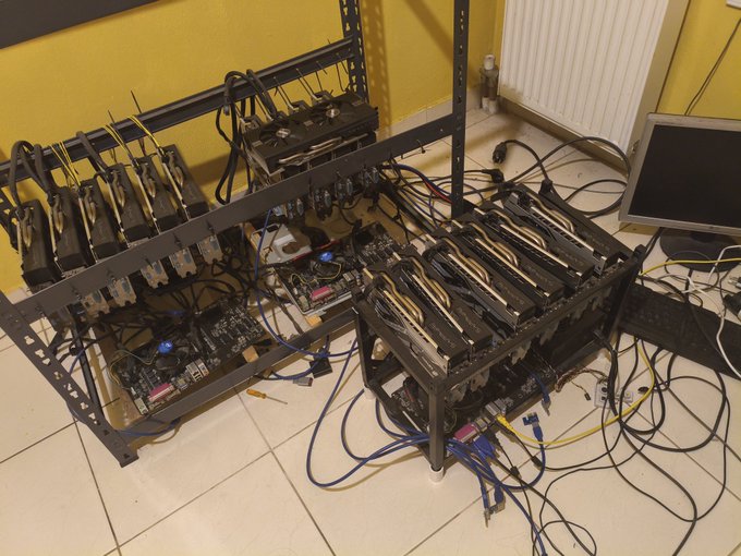 A cryptocurrency mining rig setup with multiple GPUs mounted on open-air frames, connected with various cables and a monitor displaying the operation, set up on a tile floor near a yellow wall.