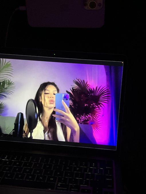 A photo of a tablet screen displaying a content creator taking a mirror selfie with a phone, with a pop filter microphone visible and purple lighting with palm leaves in the background.