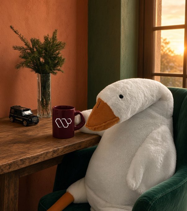 A white plush duck sits in a green armchair next to a wooden table holding a burgundy W mug, a small black car, and a vase with pine branches, lit by warm sunset light.