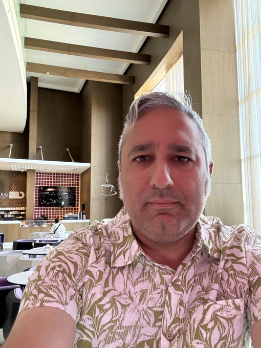 A man with graying hair wearing a pink floral Hawaiian shirt takes a selfie in what appears to be a hotel lobby or café with a Nespresso station in the background labeled 'NESPRESSO DEAR IRIS' and 'IRIS'.