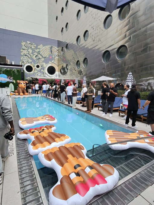 A pool party scene featuring large inflatable pool floats shaped like bundle of dynamite sticks, with people gathered around the pool and distinctive circular architecture visible on the building in the background.