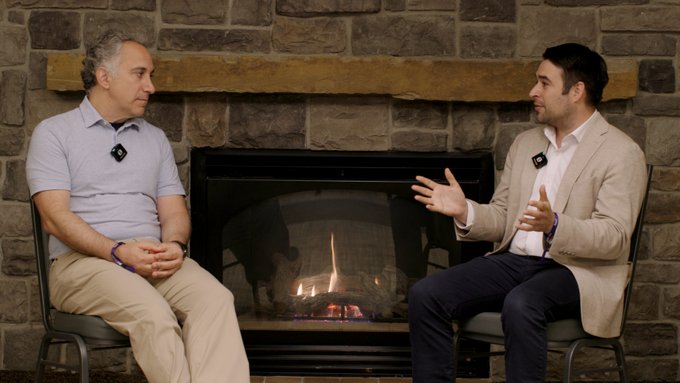 Two men seated in chairs facing each other in front of a stone fireplace, appearing to be in conversation, both wearing lapel microphones.