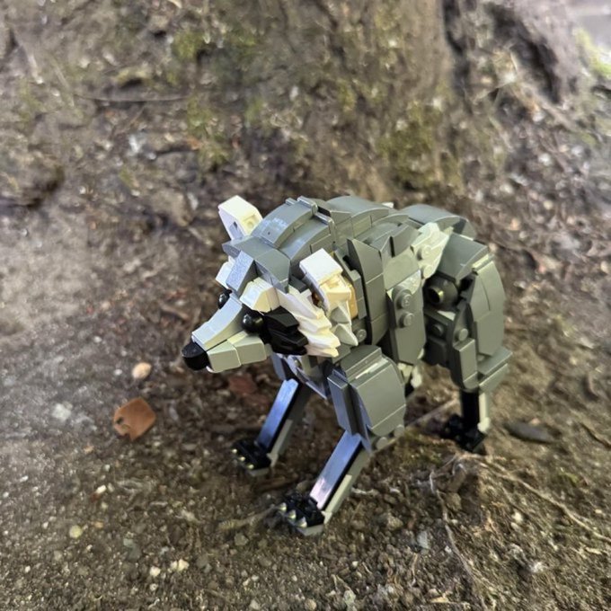 A small LEGO figure of a gray and tan wolf or dog standing on rocky, earthy ground near a tree trunk.