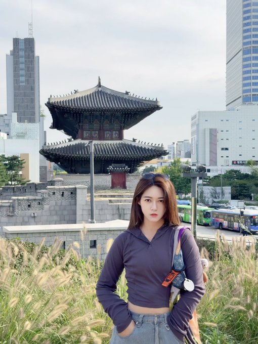 A young woman in a purple cropped hoodie and jeans stands in front of a traditional Korean palace gate and modern city skyscrapers.