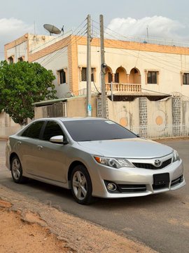 Photo of a silver Toyota Corolla sedan parked on a street in front of a building with satellite dishes on the roof, suggesting a Middle Eastern or African setting.