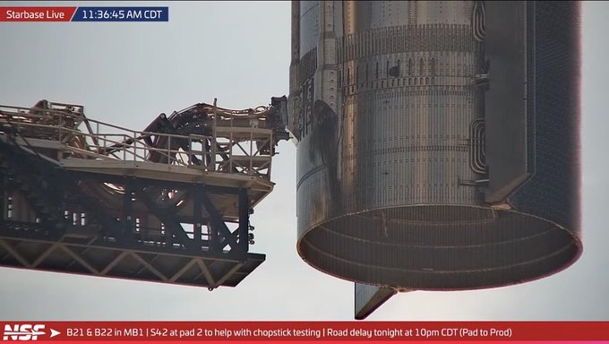 A close-up view from NSF's Starbase Live stream shows the launch tower's chopstick arms extended toward Ship 42, which is suspended on the pad, as technicians reportedly adjust the booster's height in preparation for the first chopstick catch testing, with the on-screen ticker noting "B21 & B22 in MB1 | S42 at pad 2 to help with chopstick testing | Road delay tonight at 10pm CDT (Pad to Pad)."