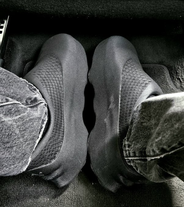 A black-and-white photo shows a pair of feet in socks and sneakers resting on a car floor mat.