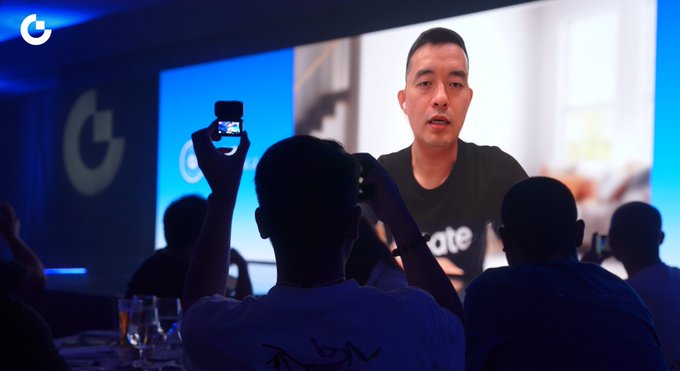 An audience member records on his phone a man displayed on a large screen at what appears to be a Gate.io branded event, where a speaker shares crypto market advice about resilience and long-term commitment across market cycles. The partially visible text "ate" suggests the speaker is associated with the Gate.io cryptocurrency exchange platform.