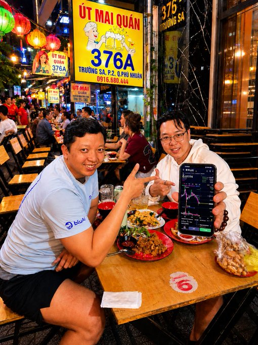 Two men sitting at a table at an outdoor Vietnamese restaurant (Út Mài Quán, 376A Trường Sa), with one holding up a phone displaying a trading chart.