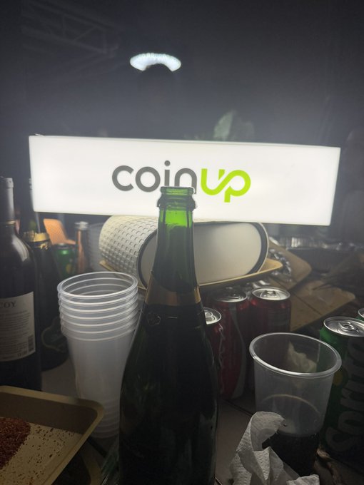 A cluttered table scene with a green glass bottle, plastic cups, soda cans, and an illuminated 'coinup' sign in the background.