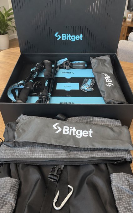 An open Bitget promotional gift box containing branded merchandise including a tripod, pouches, and accessories, alongside a branded duffel bag and backpack.