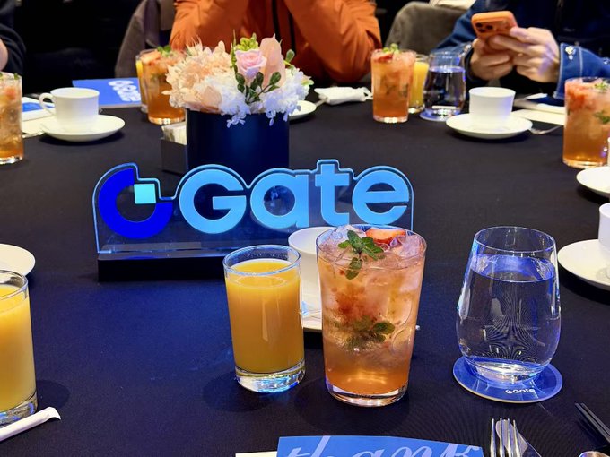A table setting with various drinks including orange juice and cocktails, centered around an illuminated 'Gate' (Gate.io) logo sign.