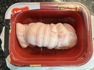 A packaged raw meat product, appearing to be a rolled and tied pork or poultry cut, sitting in a red plastic tray labeled 'HAI NGAY'.