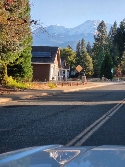 A street view showing a house with solar panels and a snow-capped mountain in the background, taken from the hood of a vehicle.