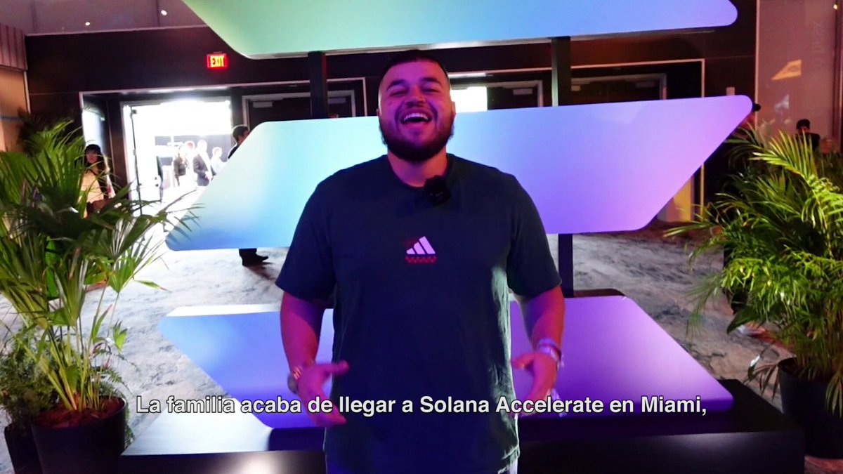 The image features a man standing next to a table with a sign that reads "Solana Accelerate Miami". The man appears to be smiling, possibly posing for a photo or giving a speech at an event. There are also potted plants visible in the scene, adding some greenery and decoration to the setting.