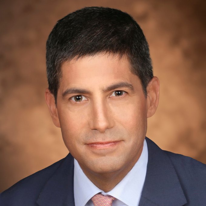 A formal portrait photograph shows a middle-aged man with dark hair wearing a dark blue suit, light blue shirt, and a pink patterned tie against a mottled brown background.
