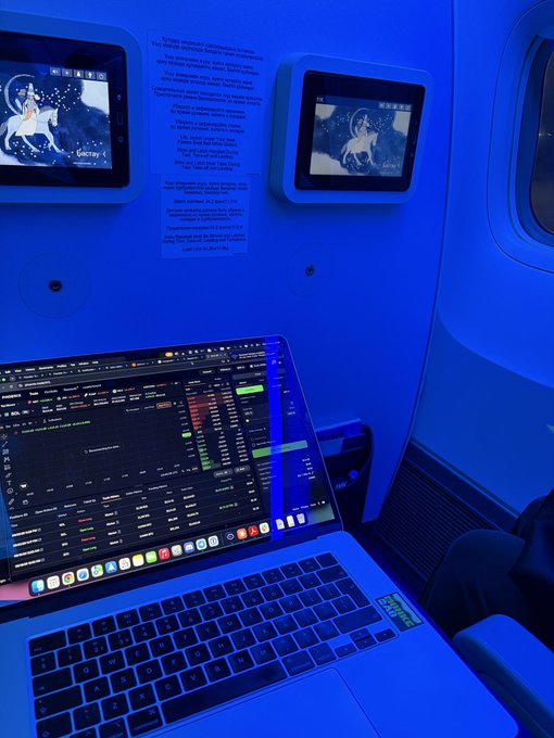 A dark blue-lit interior of an airplane showing seatback screens displaying zodiac-themed imagery, with a laptop on a tray table displaying what appears to be a cryptocurrency trading or analytics interface.
