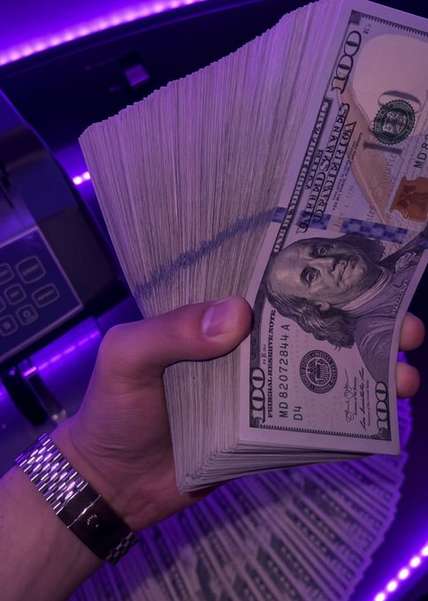 A hand wearing a metal watch band holds a thick stack of US $100 bills (serial MD 82072844 A) under purple LED lighting, with additional stacks of money and a keypad visible in the background.