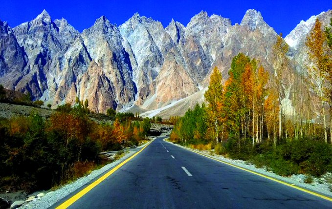 A scenic road winds through a vibrant autumn landscape in Skardu, Pakistan, framed by towering rugged mountain peaks under a deep blue sky. The image captures the breathtaking natural beauty of the region, aligning with the excitement of Binance hosting a meetup in one of Pakistan's most picturesque destinations.