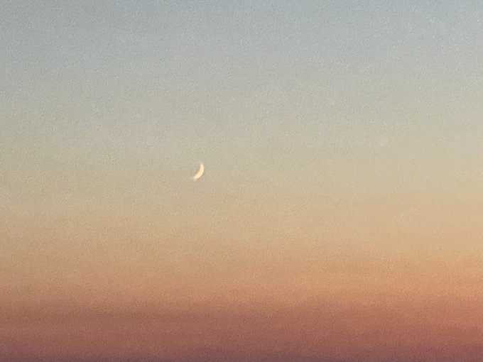 A photograph of a thin crescent moon hanging in a gradient sky transitioning from pale blue at the top to warm peach and pink tones near the horizon.