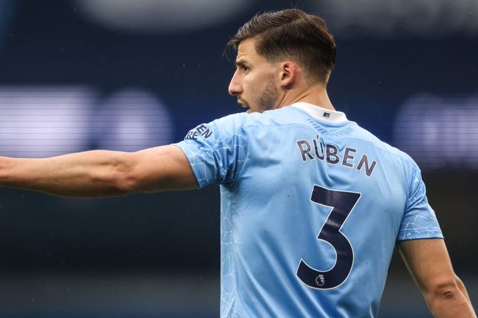 A rear view of a Manchester City football player named Ruben, wearing the light blue jersey with the number 3, pointing his arm on the pitch.