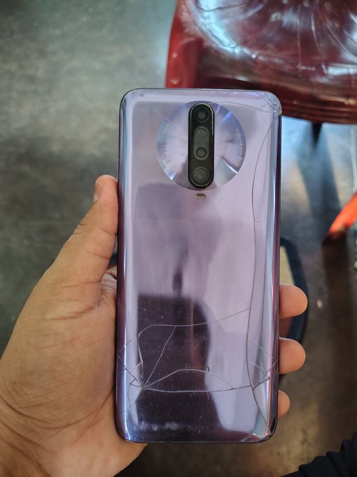 A hand holding a purple/lavender smartphone with a cracked back glass, showing a circular camera module design.