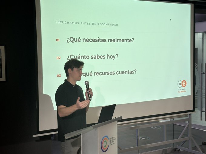 A young man presents at a podium in front of a screen displaying Spanish text: 'ESCUCHAMOS ANTES DE RECOMENDAR' with questions like '¿Qué necesitas realmente?'.