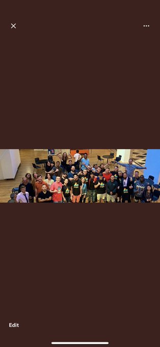 A group photo of a large crowd of people gathered in what appears to be a conference room, with some wearing dark shirts with logos.