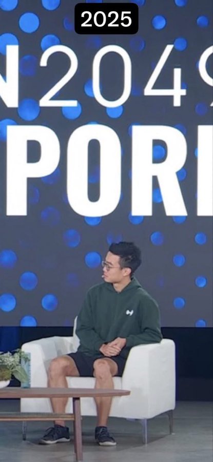 A young man in a green hoodie and dark shorts sits in a white armchair on a stage, with a large screen behind him displaying '2025' and partial text reading 'N2049 PORT'.