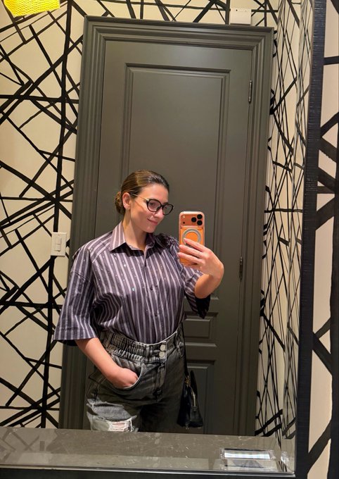 A woman wearing glasses, a striped purple shirt, and dark jeans takes a mirror selfie with an orange phone case in a bathroom with geometric black and white wallpaper.
