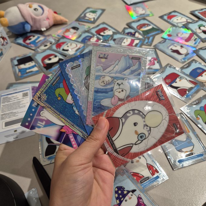 A hand holding a fan of Pokémon Trading Card Game cards, including what appears to be a Glaceon ex card and a Pachirisu card, with numerous other sleeved cards spread across a table alongside a plush toy.