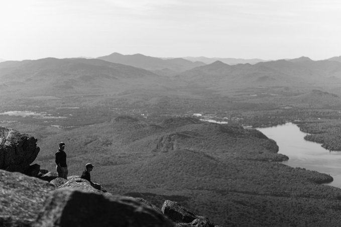 A black-and-white photo of two people standing on a rocky mountain overlook, gazing out over a vast forested valley with a winding river and distant mountain ranges.