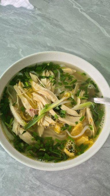 A bowl of Asian-style noodle soup with chicken, enoki mushrooms, bean sprouts, and green onions in a clear broth.
