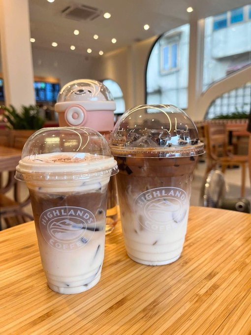 Two iced coffee drinks from Highlands Coffee sit on a wooden table in a bright café, one in a pink dome-lidded cup with a child-sized pink water bottle in the background.