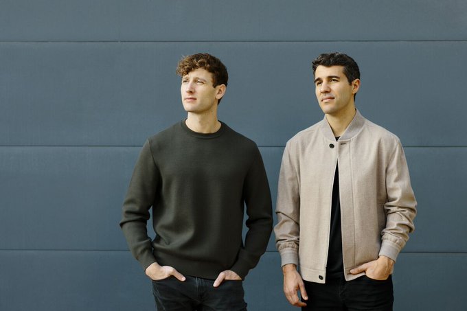 Two men stand side-by-side against a blue paneled wall, both gazing upward—the left man in a black sweater with hands in pockets, and the right man in a beige bomber jacket. The contemplative, forward-looking pose visually echoes the context about Harvey, the legal AI startup reportedly facing mounting financial strain as reliance on rented OpenAI and Anthropic model capacity drove gross margins sharply negative.