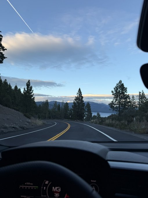 A view from inside a car driving along a winding road through a mountainous, tree-lined landscape under a partly cloudy sky at dusk, with the dashboard display showing "M3" and "35" alongside a speedometer. The scenic drive perfectly complements the relaxed "Not bad" sentiment.