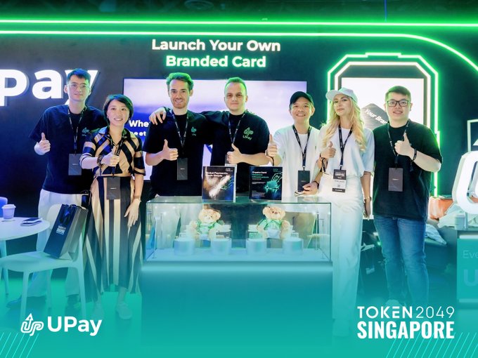 A group of seven people posing with thumbs up at a TOKEN2049 Singapore event for UPay, with the banner 'Launch Your Own Branded Card' displayed behind them and plush mascots in a display case.