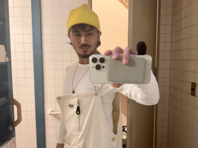 Mirror selfie of a young man wearing a yellow beanie, white long-sleeve shirt, and cream overalls, holding an iPhone with a light green case in a tiled bathroom.