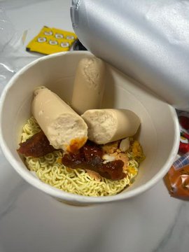 An unconventional food combination in a white paper cup, featuring instant noodles topped with what appears to be sausages and dark sauce.