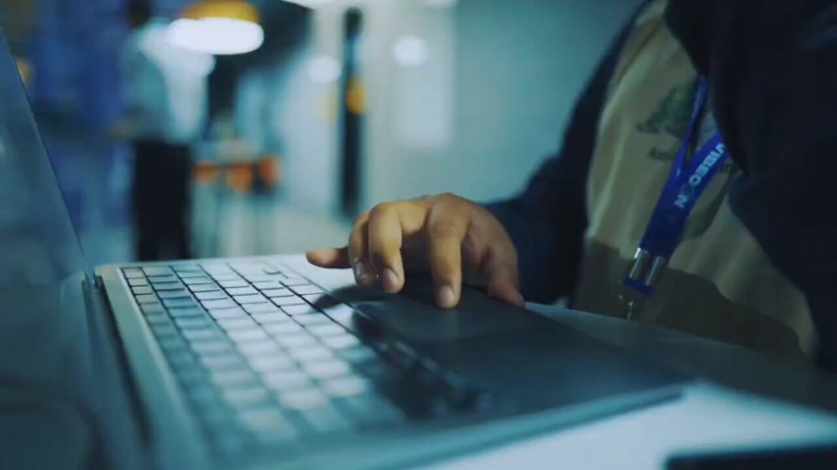 The image shows a person's hand typing on a laptop computer with their fingers moving across the keyboard. The background appears blurry, but there are some people visible behind the laptop screen. The focus of the image is on the action of typing, capturing the essence of someone working or creating content using technology.