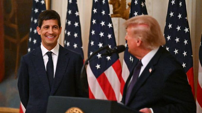 The image shows two men in suits standing in front of American flags, with one man speaking at a podium equipped with a microphone. Both are dressed in formal dark suits with ties, and the setting appears to be an official government or ceremonial event. The accompanying post discusses a mysterious $122 million cryptocurrency trade made just before the Federal Reserve's 2 p.m. decision, though the image itself does not directly depict this trade or any Fed-related event, instead showing what appears to be a political or governmental ceremony.