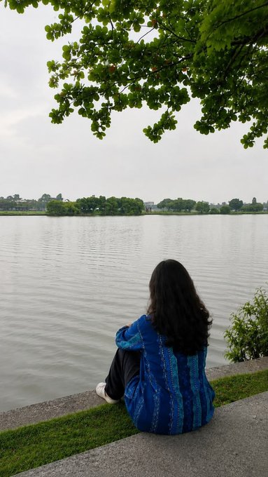 A woman with long dark hair wearing a blue patterned outfit sitting on a concrete ledge by a lake, looking out at the water with trees framing the scene.