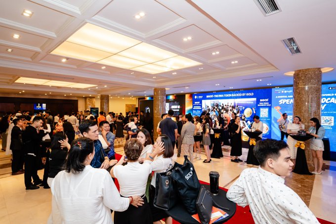 A crowded indoor networking event or conference with many attendees gathered in a hotel lobby area, featuring digital screens displaying 'SENO - THE FEEL YOUNG BACKED BY GOLD' and other promotional content.