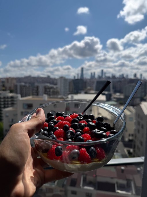 A hand holding a glass bowl filled with raspberries, blueberries, and boba pearls against a backdrop of a city skyline under a cloudy blue sky.
