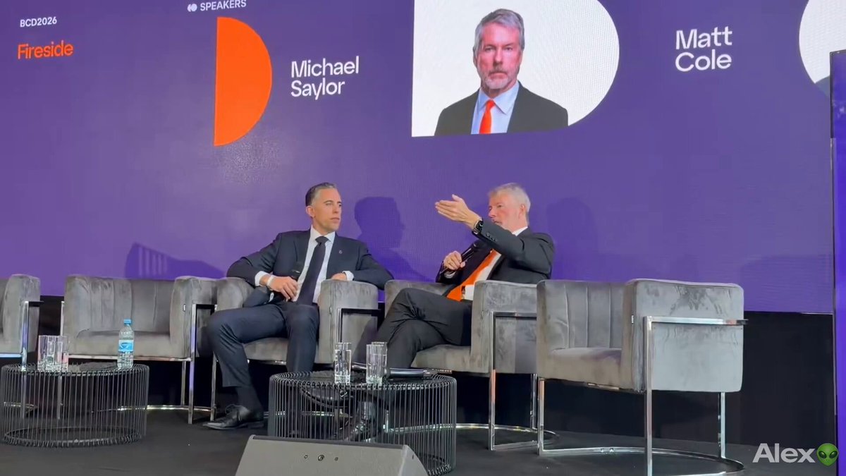 The image shows two men sitting in chairs at a conference room with a purple wall behind them. One man has a tie visible while the other one does not. They are both facing each other, engaged in conversation. The background features an orange sign that reads "FREESIDE" and another sign that says "MATT COLE". There is also a microphone positioned near the men, suggesting they might be participating in a panel discussion or presentation.